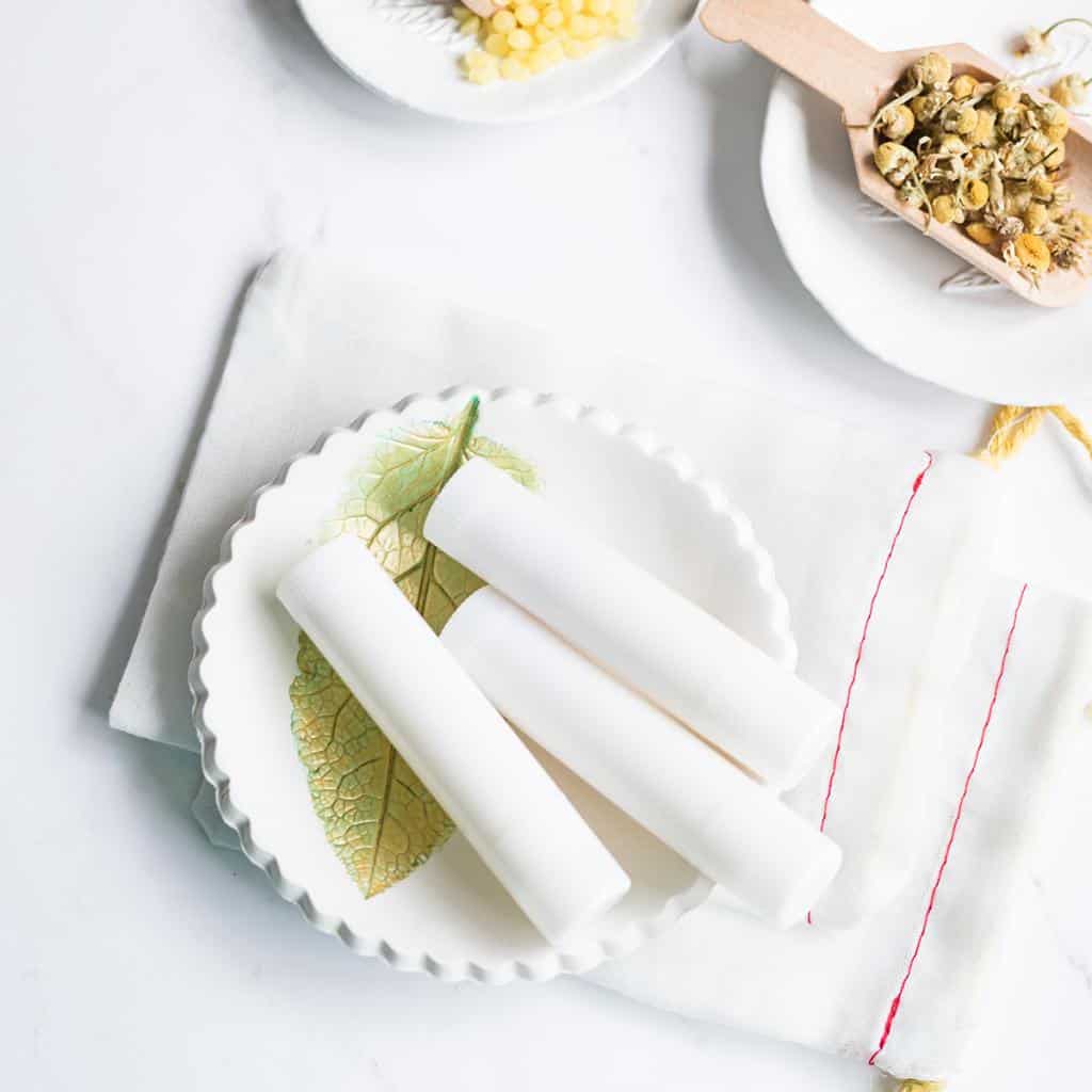 creamy smooth lemon chamomile lip balm recipe inside white lip balm tubes on a handmade clay bowl next to supplies.