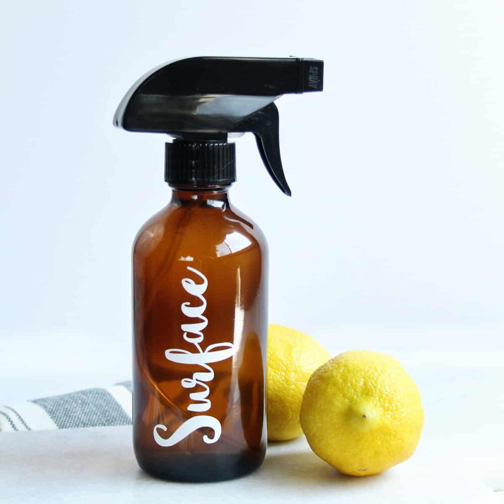 amber glass cleaning bottle labeled surface on a white countertop next to bright yellow lemons and a cleaning towel