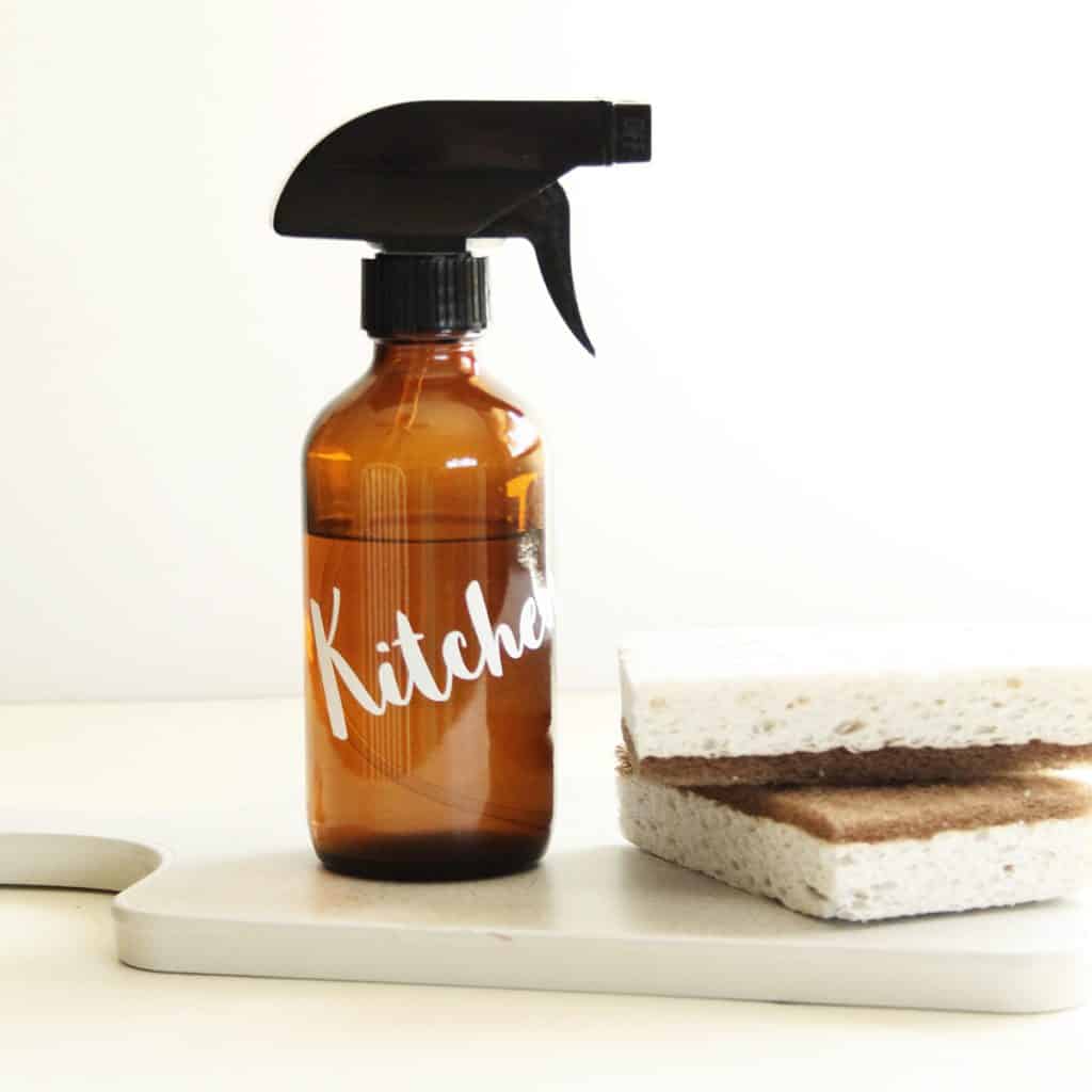 amber glass spray bottle with the word ktichen on a cutting board next to a stack of cleaning sponges.