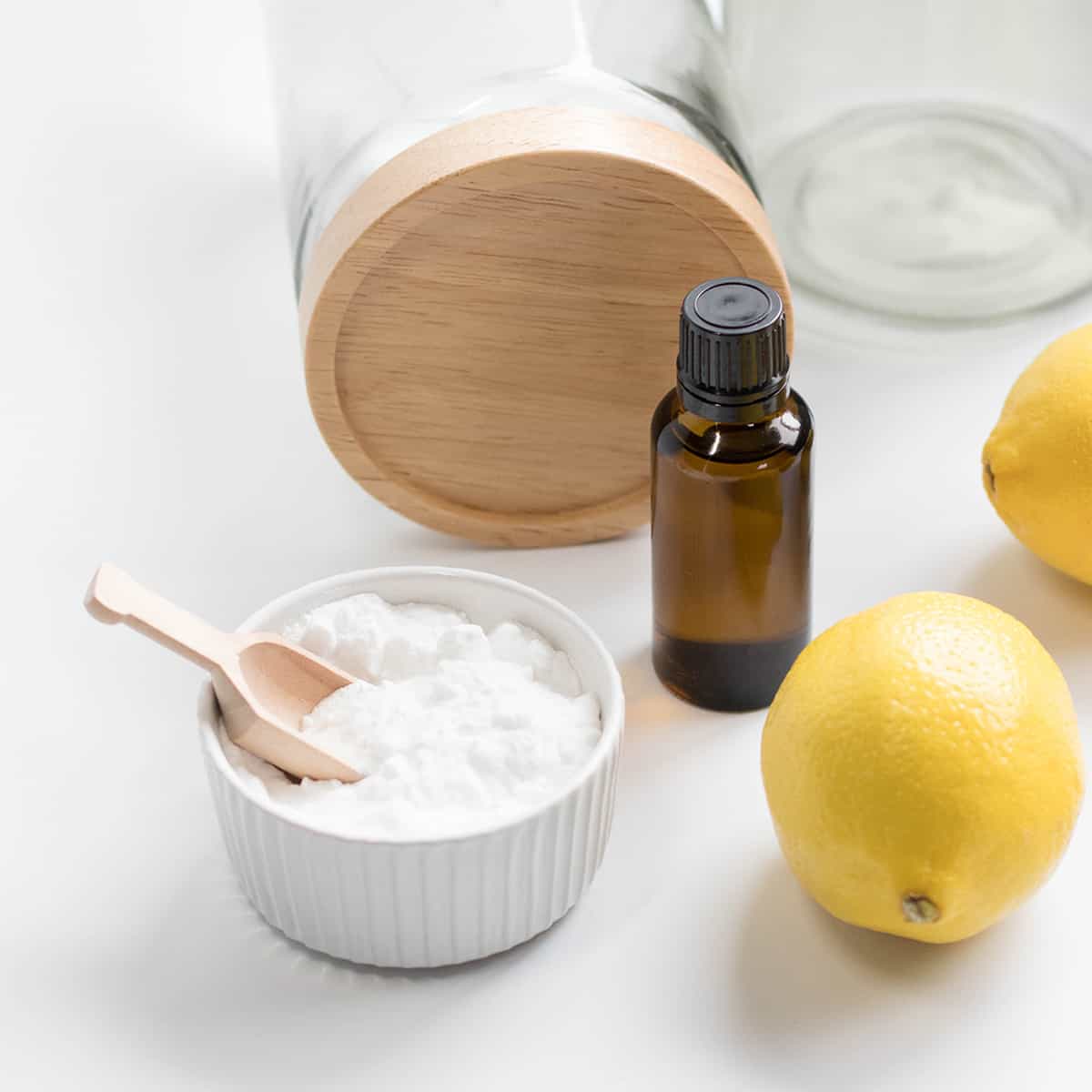 A small white bowl filled with baking soda, next to fresh lemon, and pantry jars, on a bright white kitchen counter.