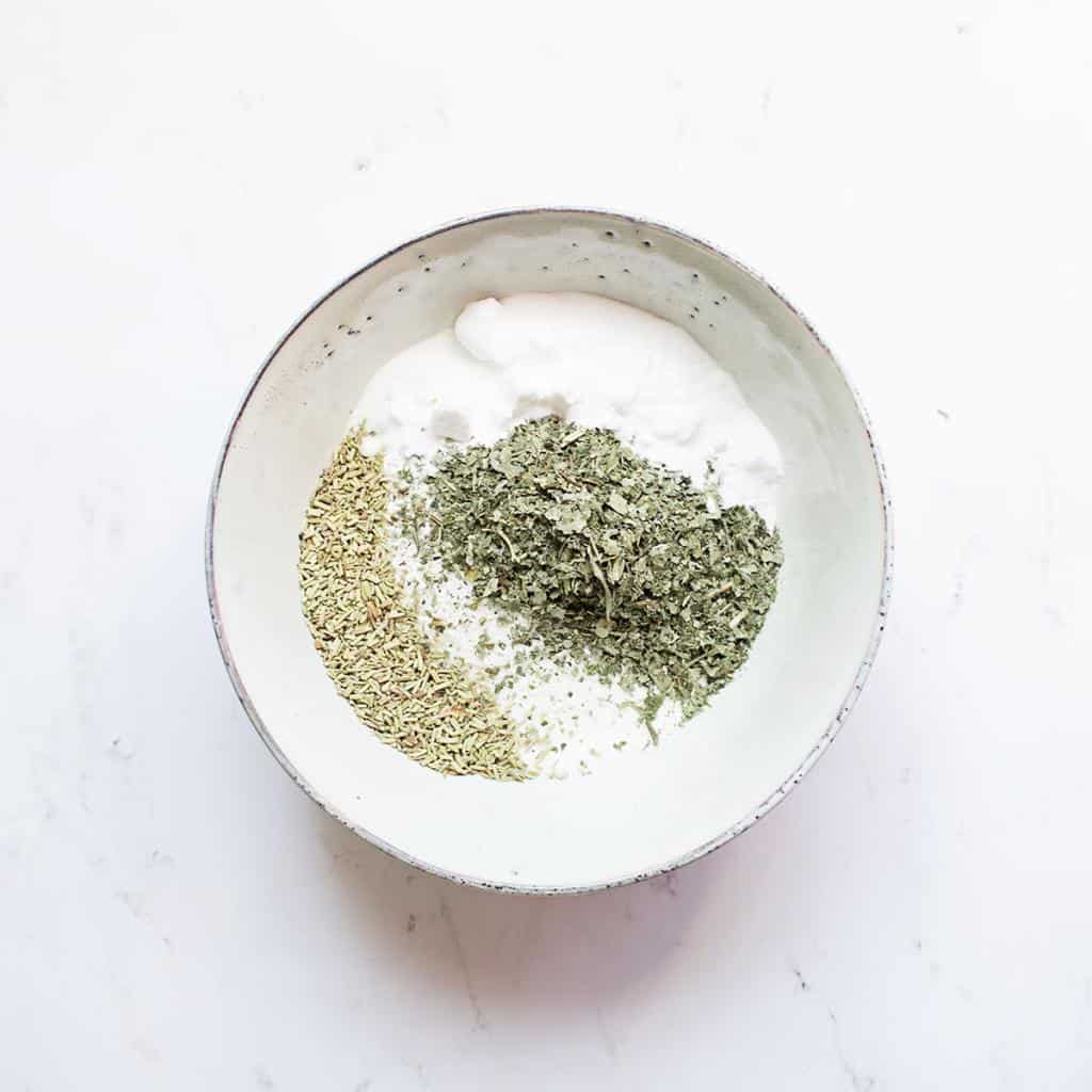 Minimalist style ceramic bowl of dried herbs, and powders for natural cleaning on a marble countertop.