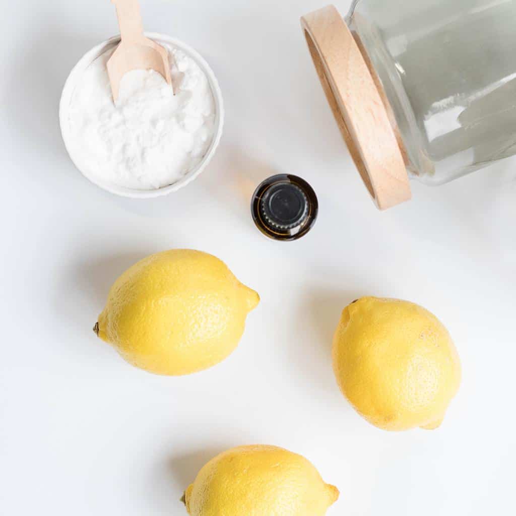 natural cleaning ingredients and supplies: glass storage jars, essential oil bottle, fresh lemons, bowl of baking soda.