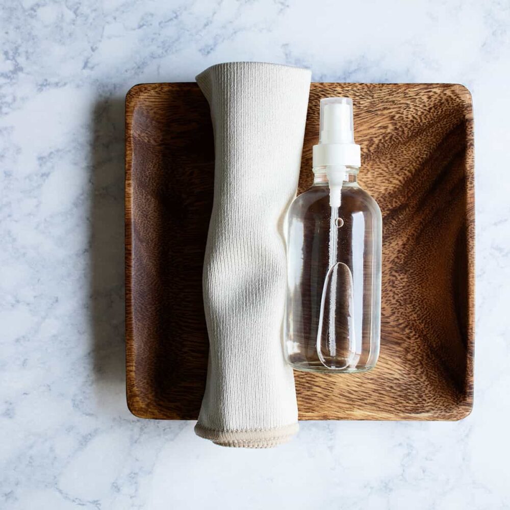 small clear glass bottle of DIY stainless steel spray on a wooden tray next to a small microfiber cloth