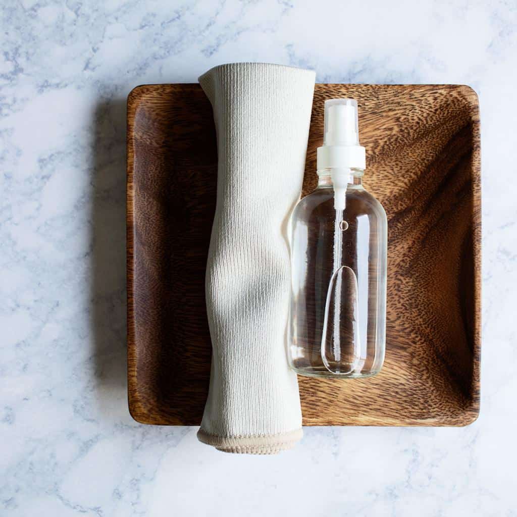 small clear glass bottle of DIY stainless steel spray on a wooden tray next to a small microfiber cloth