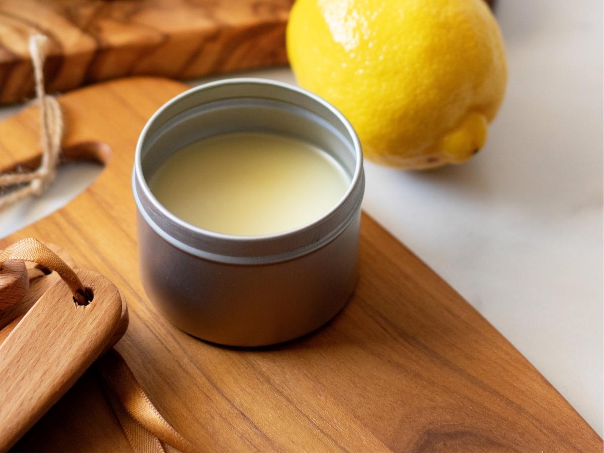 A metal tin of yellow lemon conditioning spoon butter on a wooden cutting board next to a lemon