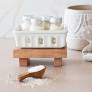 Jars of oatmeal bath ingredients in a white ceramic basket on a wood tray, behind a scoop of a oatmeal bath mix.