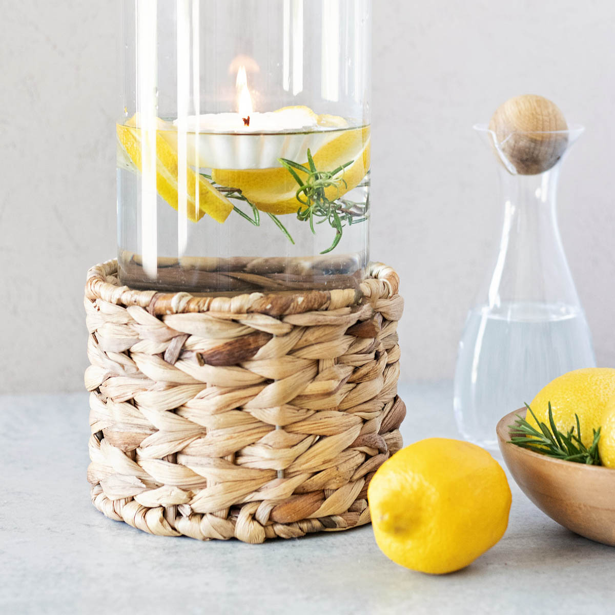 Tall clear glass vase in a rattan holder filled with fresh green rosemary, yellow lemon slices, and a white floating candle tart with a glowing candle wick.