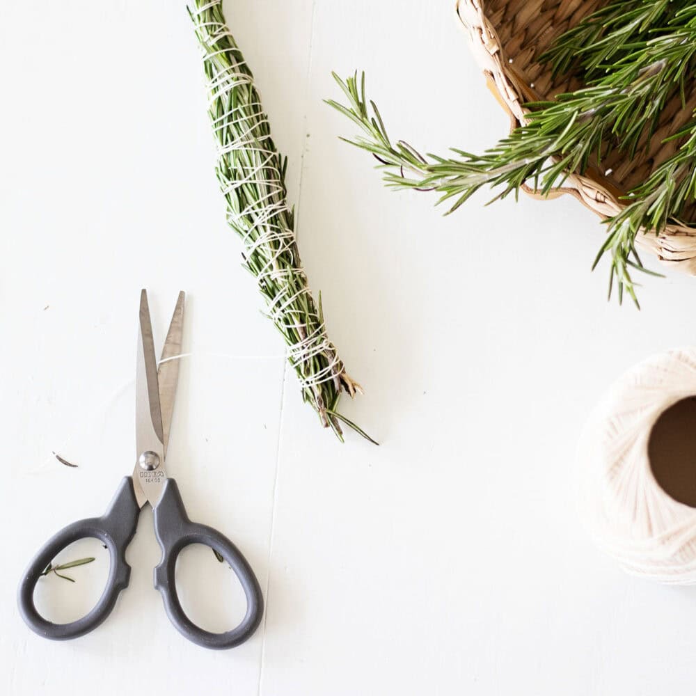 A handmade rosemary smudge stick tied with cotton twine sitting next to scissors and a basket of fresh rosemary branches