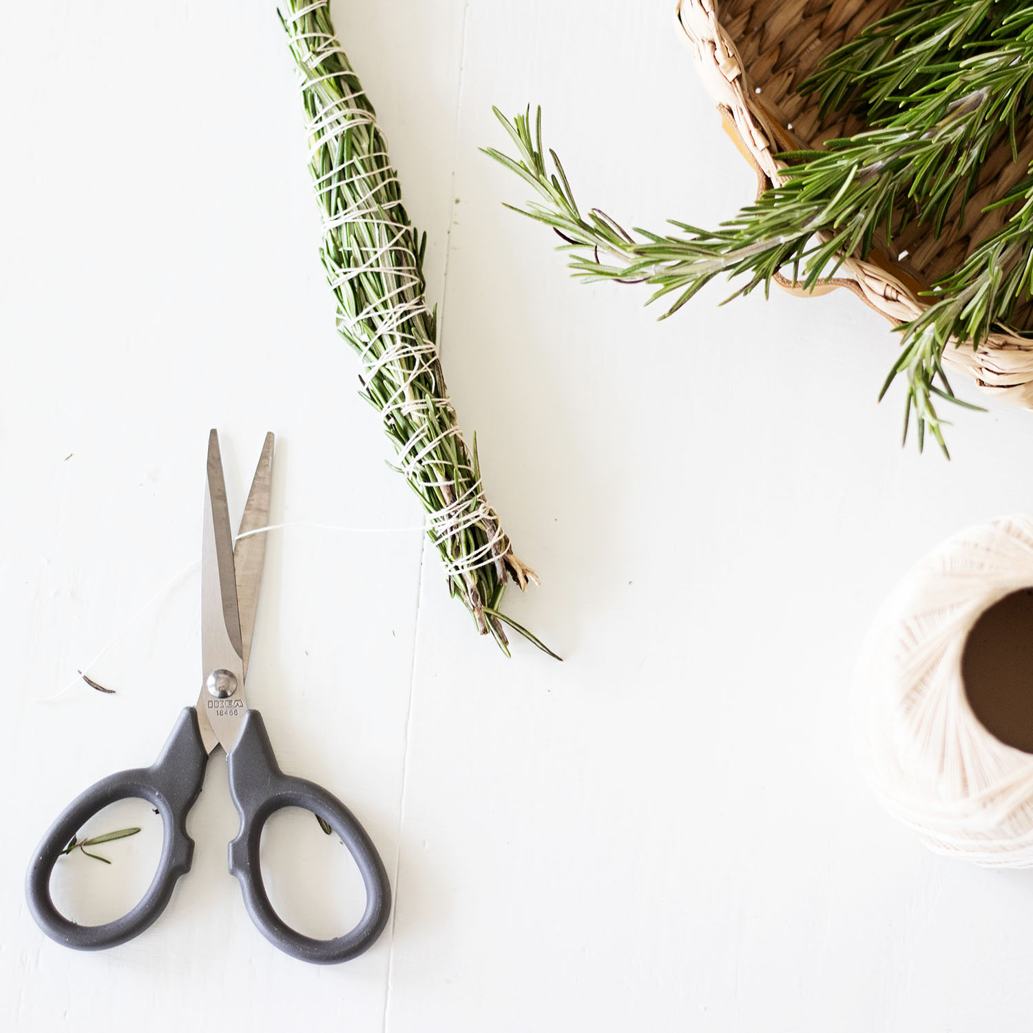A handmade rosemary smudge stick tied with cotton twine sitting next to scissors and a basket of fresh rosemary branches