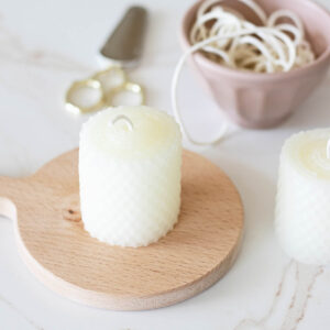 Creamy white, handmade votive candles on a marble countertop and small wood board next to scissors and a cotton wick roll.