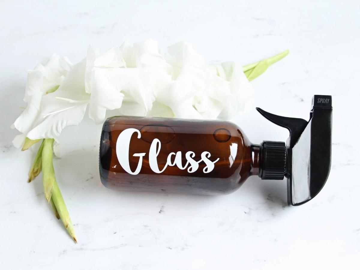 amber glass bottle labeled glass, filled with natural streak free glass cleaner, lying on a bright marble counter next to white flowers