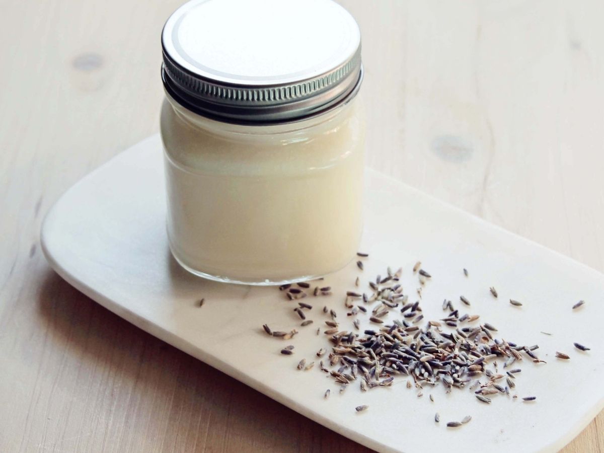 Fluffy, whipped white body butter in a glass jar surrounded by dried lavender buds on bright background.