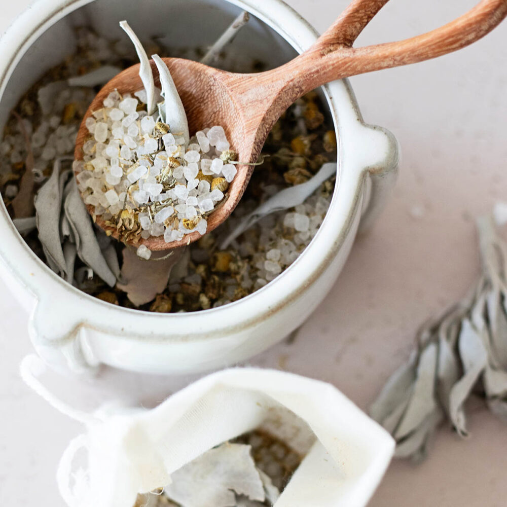 handmade soothing herbal bath soak with a spoon full of bath salts, sage, and chamomile in a small bowl next to a linen bath tea bath