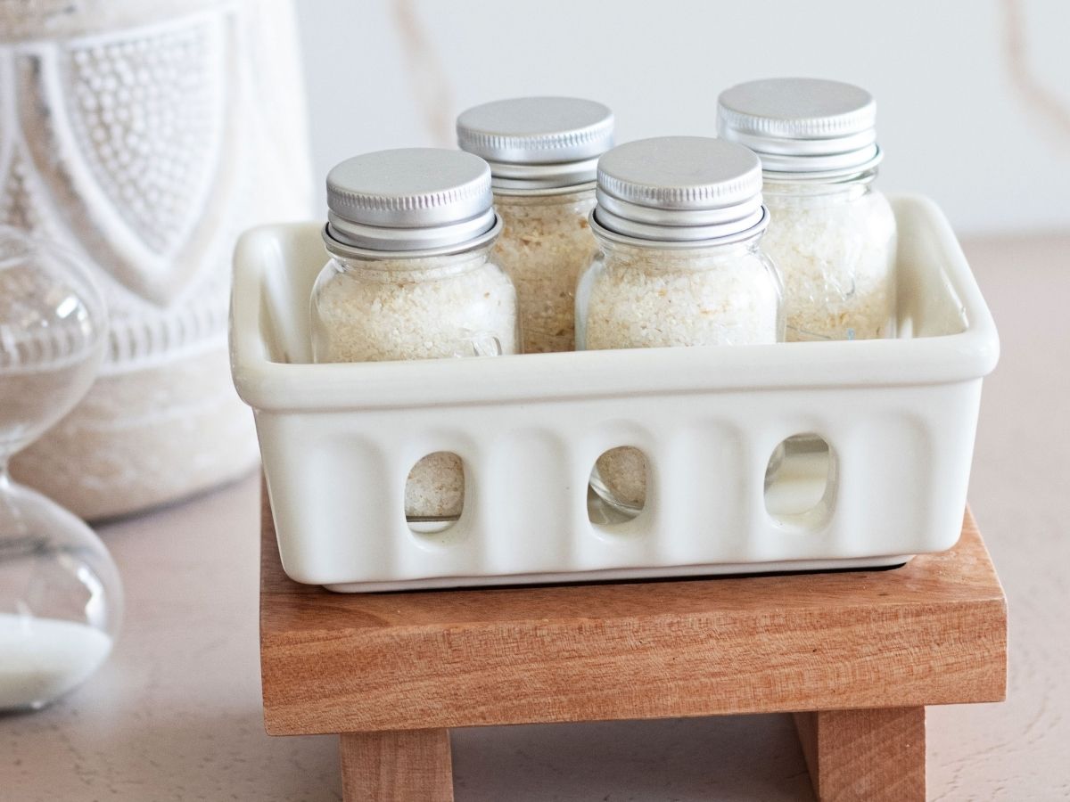 Jars of oatmeal bath ingredients in a white ceramic basket on a wood tray, behind a scoop of a oatmeal bath mix.