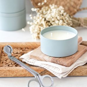 Double wick candle in a sage blue candle tin ontop of wood boards next to a candle wick trimmer.