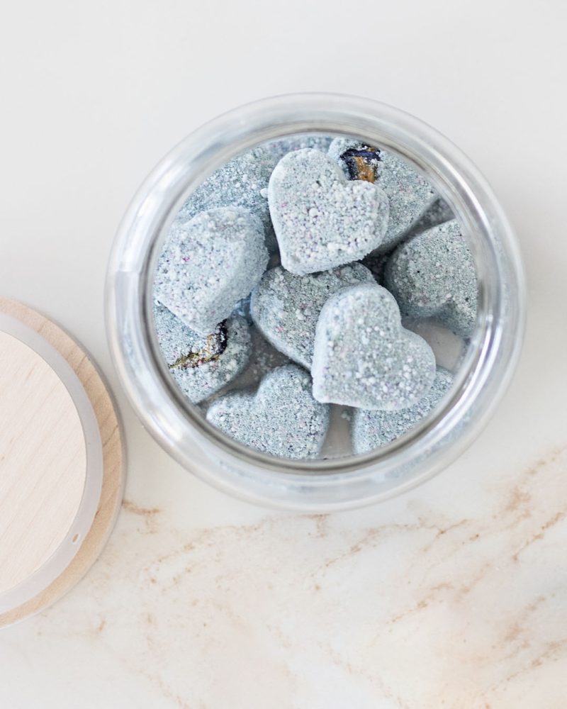 Open glass jar filled with heart-shaped blue color bath tablets on a creamy marble countertop.
