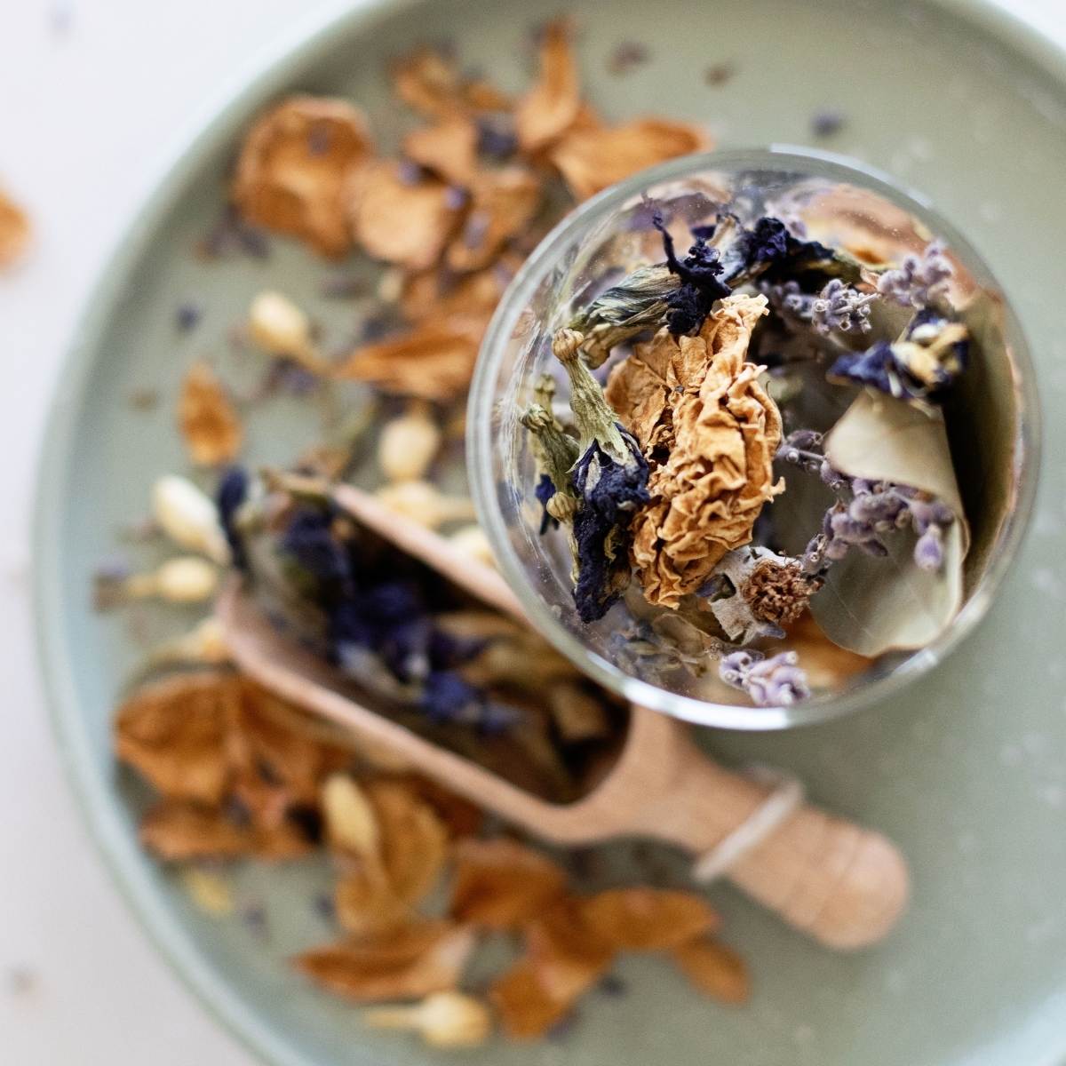Closeup looking into a big jar of botanical simmer pot ingredients on a green tray with a wooden scoop and scattered colorful petals.