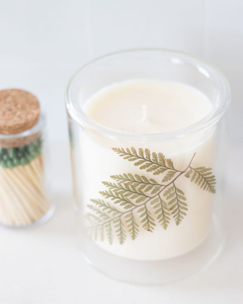 White candle in a glass jar with a real pressed leaf next to a small match jar.