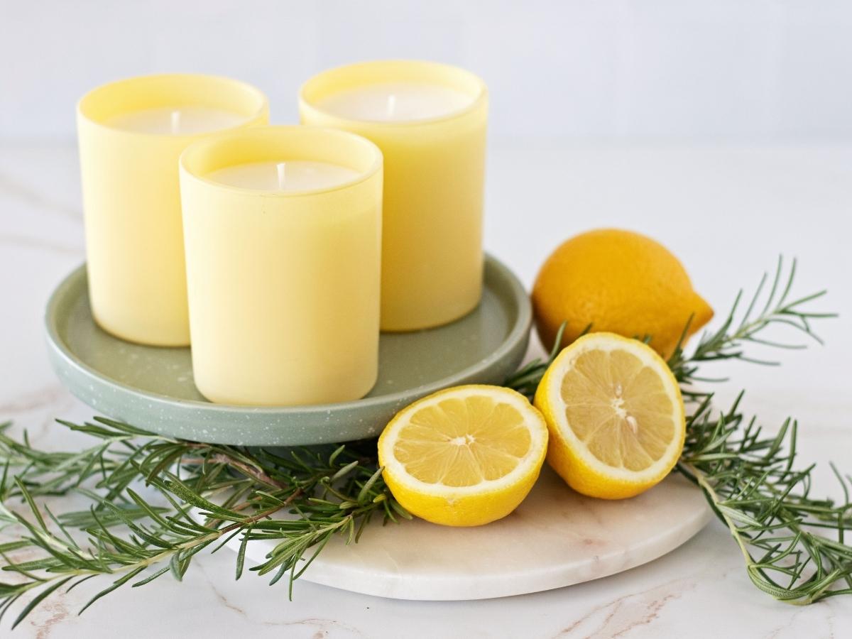 A trio handmade candles in tall sunny yellow candle jars on a sage green tray next to fresh rosemary and sliced lemons halves.