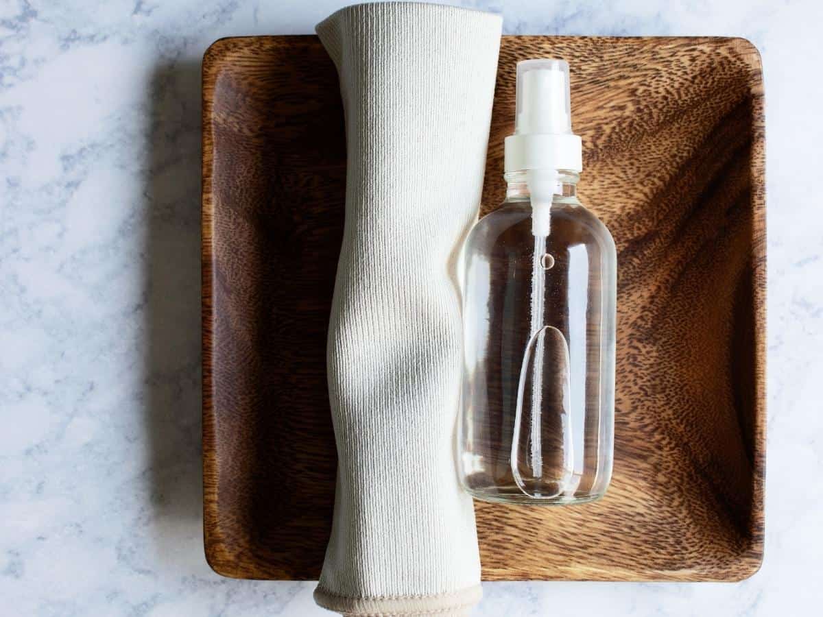 small clear glass bottle of DIY stainless steel spray on a wooden tray next to a small microfiber cloth