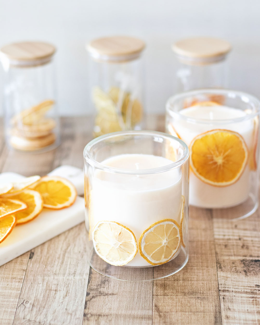 wooden table with handmade soy candles in glass jars with dried citrus inside, in front of containers of dried citrus fruits