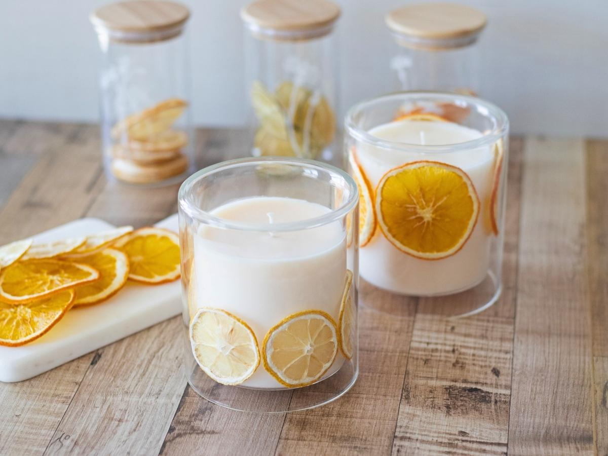 handmade soy candles in glass jars with dried citrus inside, in front of containers of dried citrus fruits on a wood table