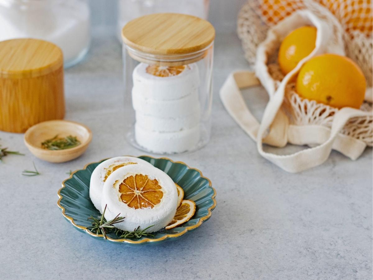 large white shower steamers filled with a whole dried orange slice and rosemary on a dark teal ruffled bowl in front of glass jar of shower steamers.