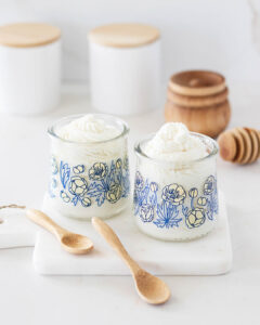 glass yogurt jars featuring a blue floral pattern filled with creamy, white whipped soap, next to mini wooden spoons on a bathroom marble counter