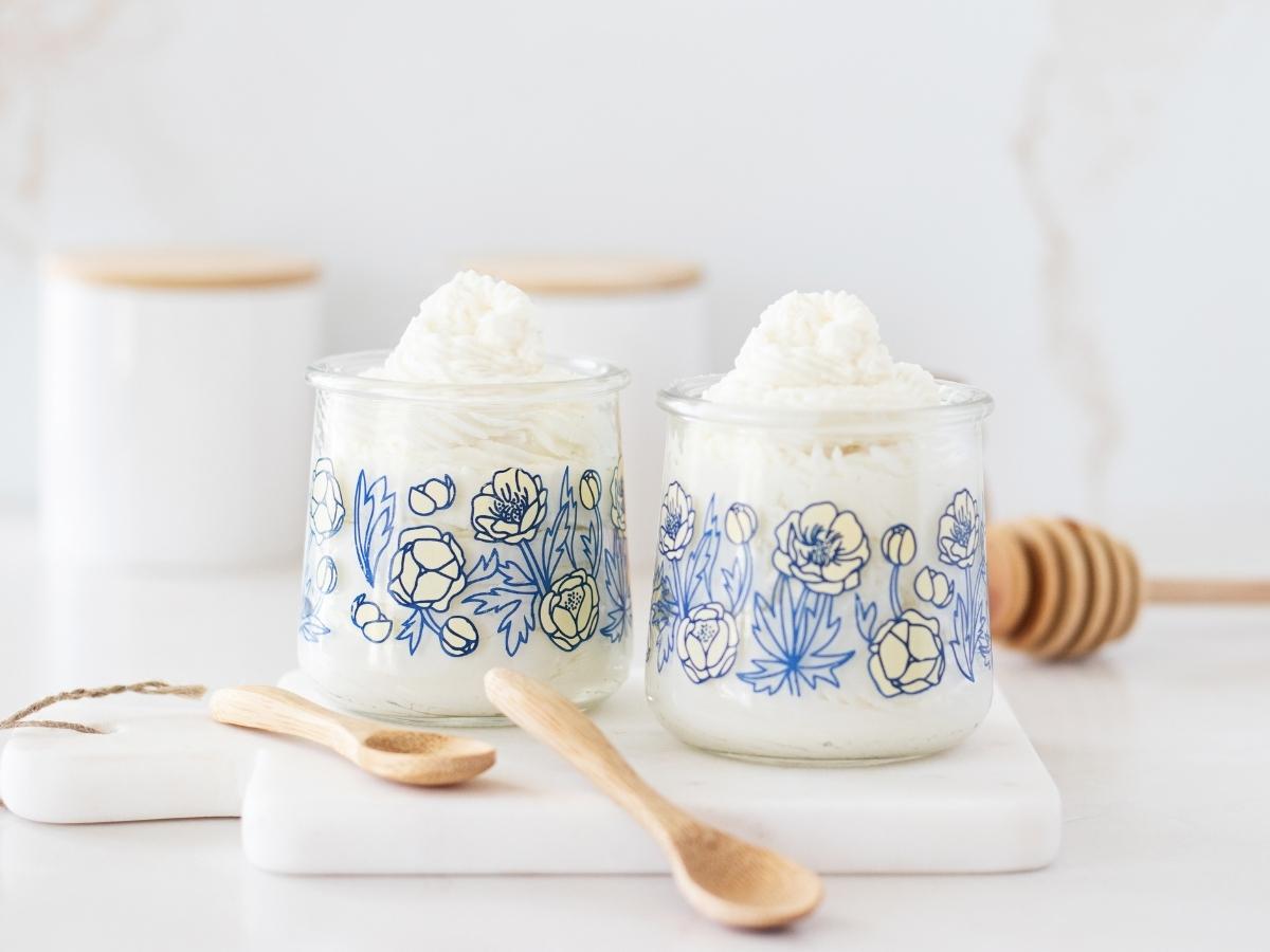 glass yogurt jars featuring a blue floral pattern filled with creamy, white whipped soap, next to mini wooden spoons on a bathroom marble counter