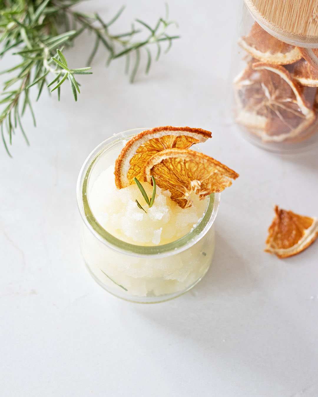 glass yogurt jar filled with a DIY hand scrub, topped with dried orange slices on a kitchen countertop next to a jar of orange slices and fresh rosemary branches.