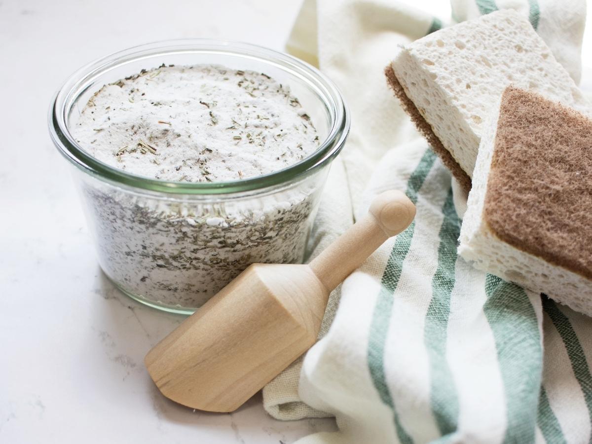 glass Welk jar of homemade scouring powder on a kitchen countertop with wooden scoop, kitchen towel, and sponges.