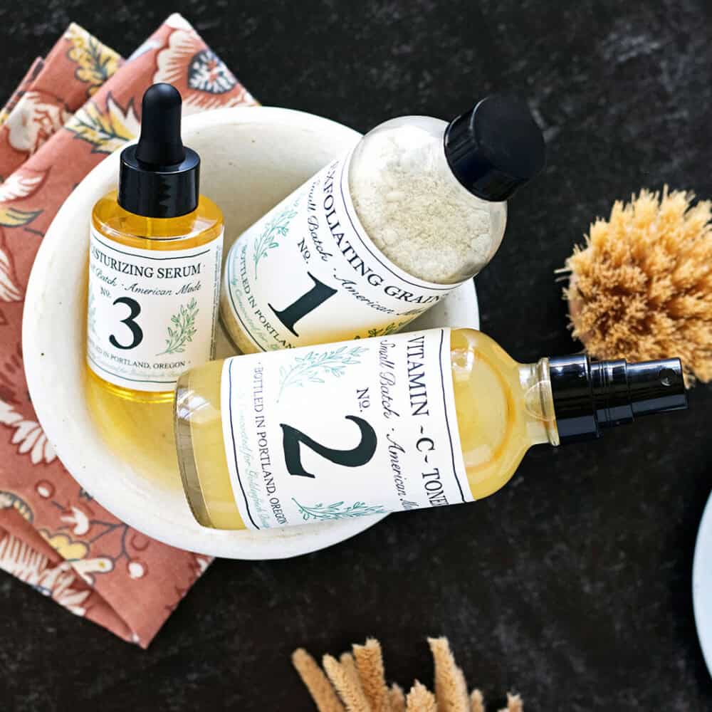 organic stone bowl of Goldenfinch Boutique skin care products on a colorful linen cloth and black marble counter next to a natural facial brush.