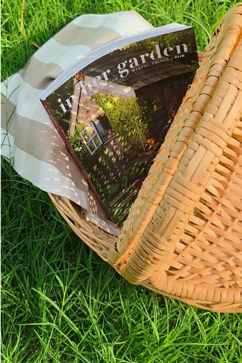 In Her Garden Magazine inside a wicker picnic basket on a green lawn.