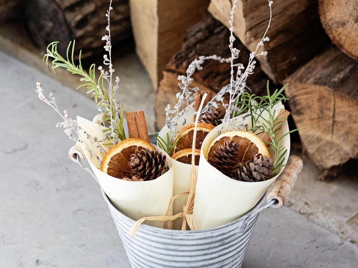 homemade fire starter bundles filled with pine cones, dried orange slices, spices, and herbs in a metal bucket in front of logs in an outdoor fireplace