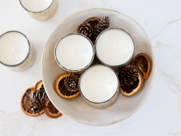 looking over a light marble countertop with two soy wax candle jars, pine cones, dried orange slices, and on the counter and large bowl filled with 3 more candles and whole spices
