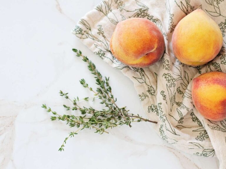 Clean marble kitchen countertop styled with fresh peaches, thyme sprigs, and a linen tea towel.